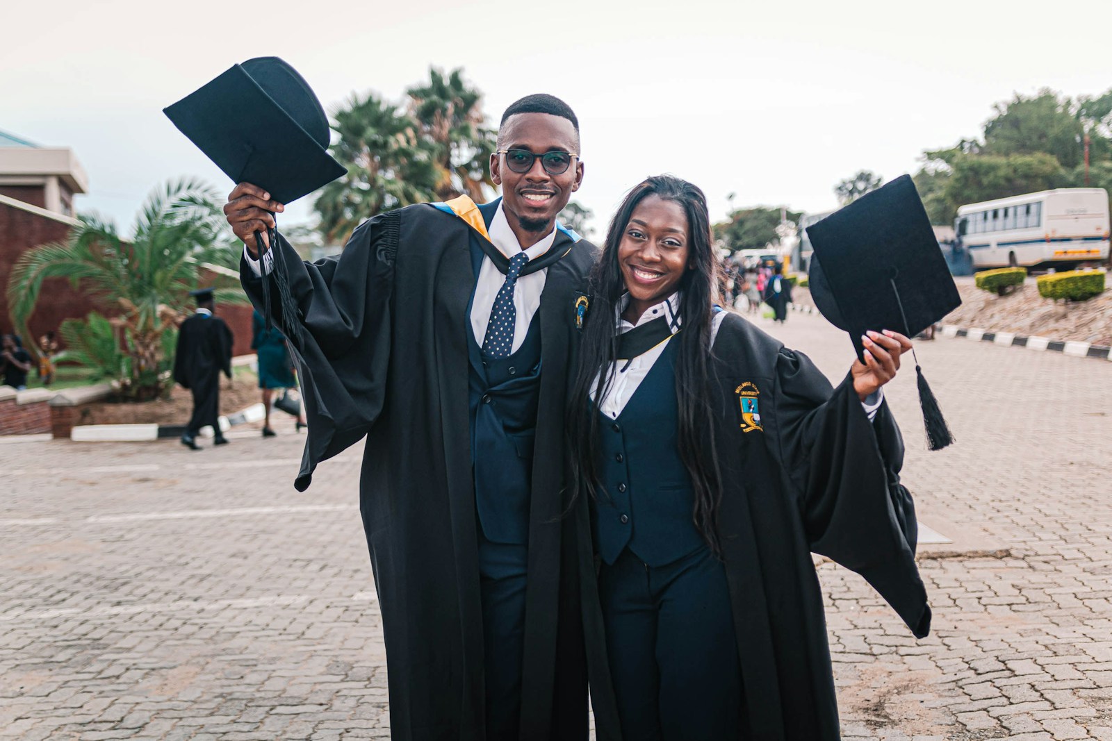 Two graduates in academic gowns raising their caps in celebration on a Ugandan campus.